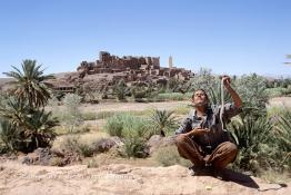 Image du Maroc Professionnelle de  Le paysage, formé par la kasbah Tifoultoute qui se dresse majestueusement sur un éperon rocheux qui domine la vallée de l'oued Ouarzazate, attire les touristes qui prennent le temps de s'arrêter pour prendre une photo. Précisément à cet endroit un charmeur de serpent tenant une vipère avec sa main gauche tout en surveillant le mouvement du scorpion noir qu’il avait accroché à son cou avec sa main droite pose aux touristes contre un billet avec derrière lui comme décor celui qui avait servit pour de grand films comme Lawrence d’Arabie et Jésus de Nazareth à quelque kilomètres de la ville de Ouarzazate au sud du Maroc le Samedi 23 Août 1997. (Photo / Abdeljalil Bounhar)
 